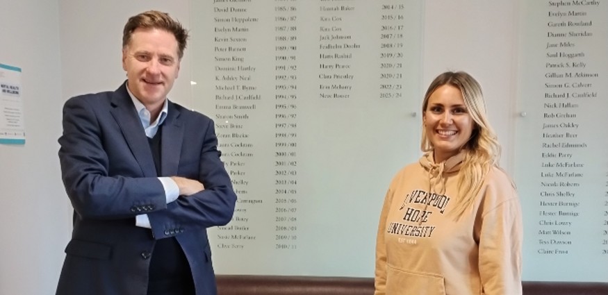 A wearing a suit and a woman wearing a hoodie stand in front of a roll of honour board.